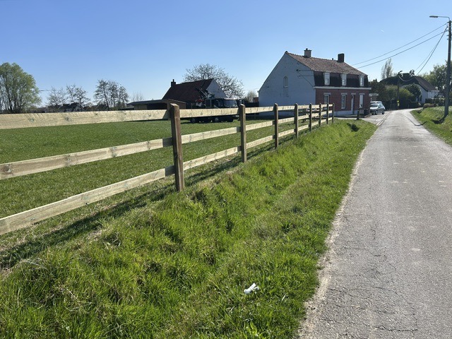 Houten omheining met drie planken langs een landelijke weg bij een groene wei en huizen