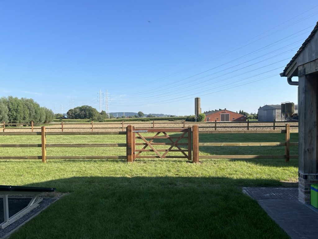 Houten omheining met poort op een groene weide onder een heldere blauwe hemel bij een boerderij