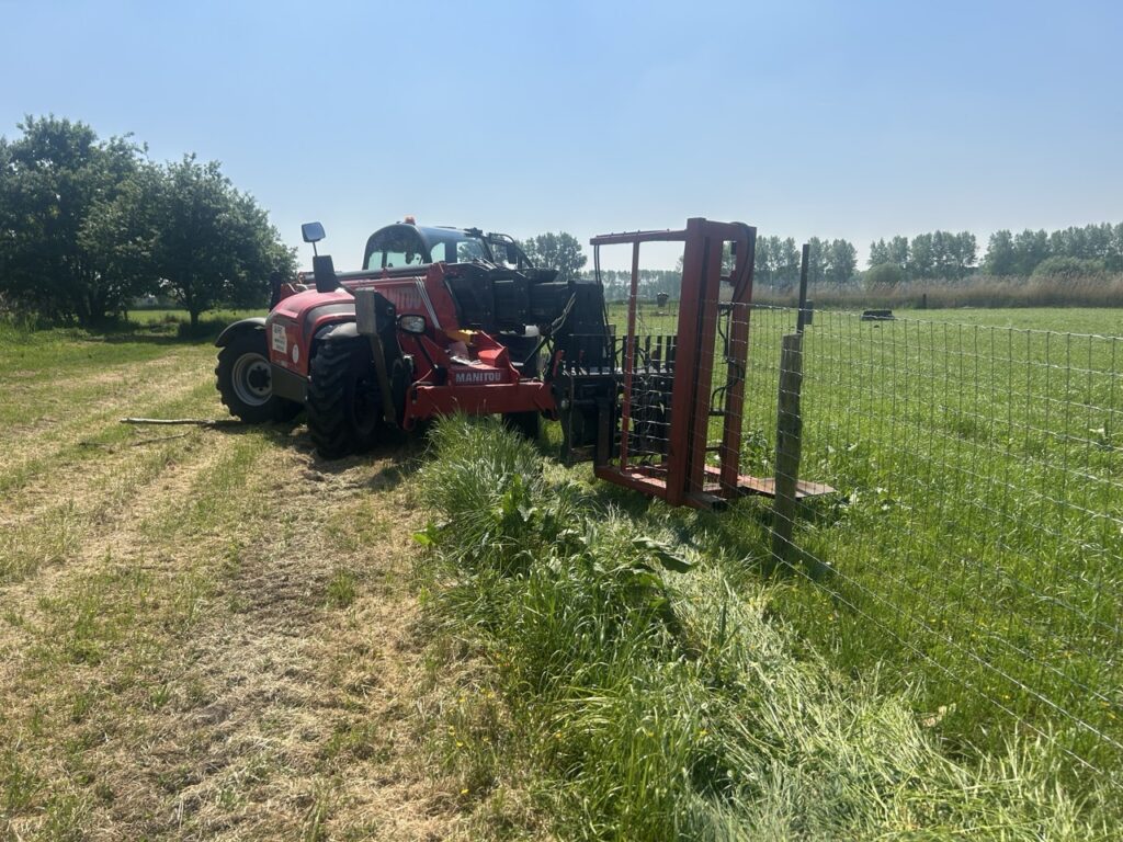 Rode landbouwmachine plaatst metalen gaasomheining in groene wei bij zonnige dag voor veedierenafbakening