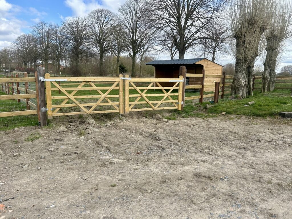 houten poort en afrastering met metalen gaas op weiland met schuilstal en bomen op achtergrond