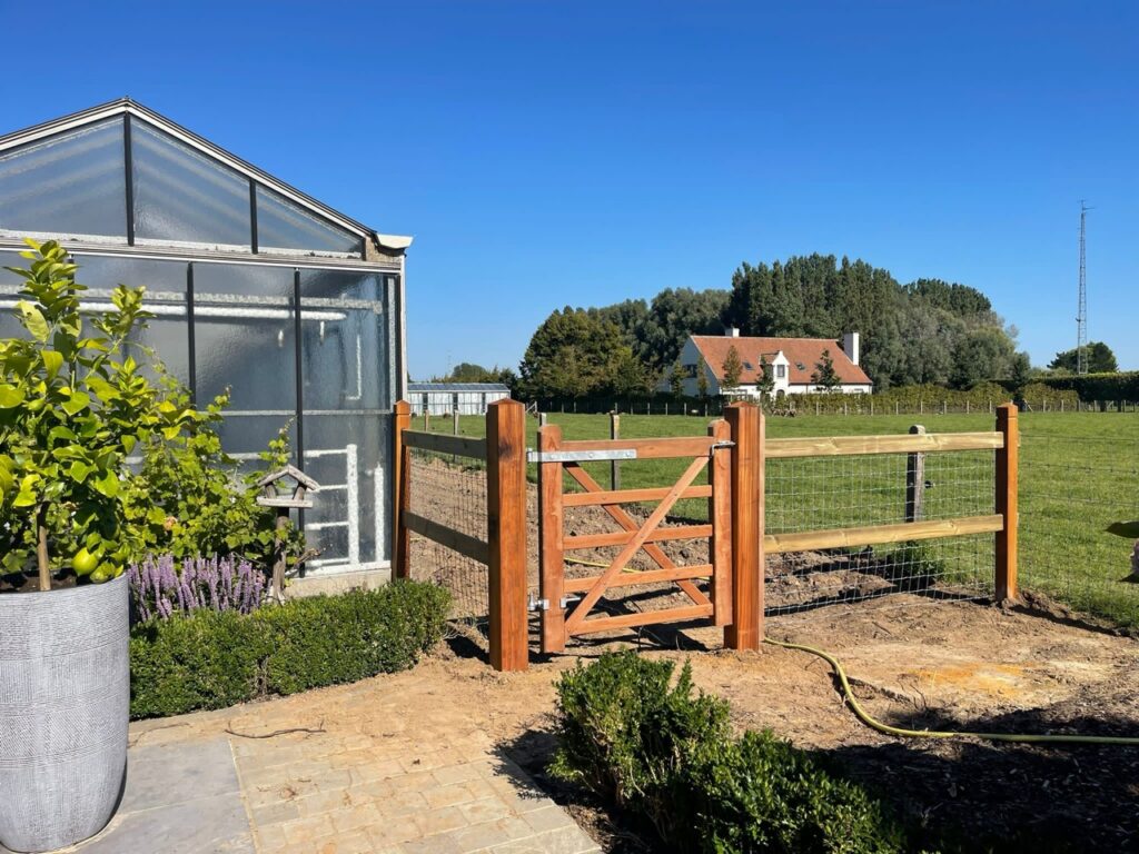 Houten tuinpoort en afrastering met gaas naast een kas onder een blauwe lucht voor dierenafscheiding