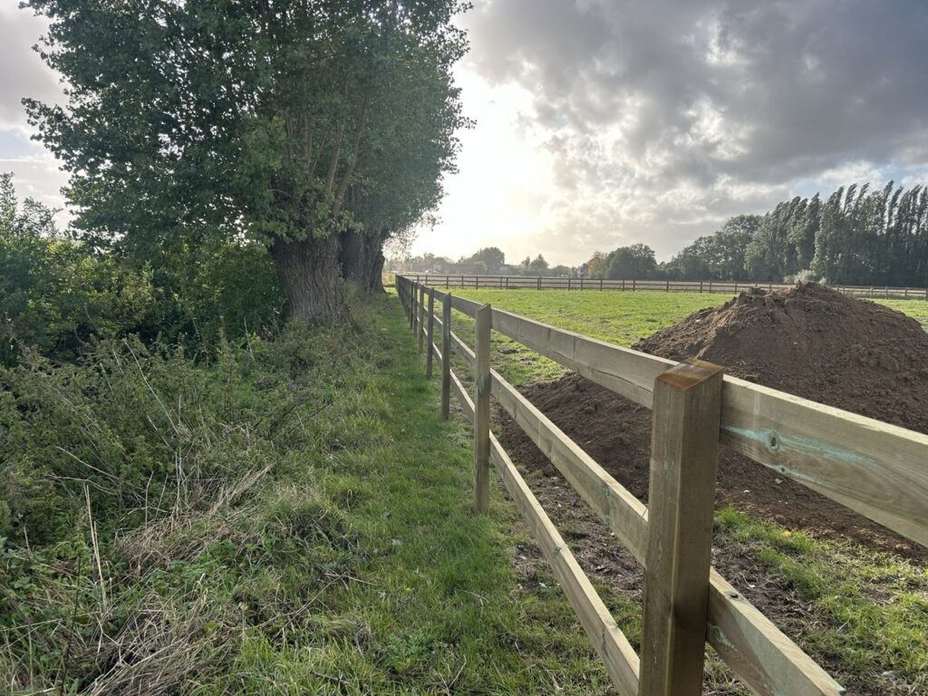 houten afrastering langs weiland met bomen en zicht op open veld onder bewolkte lucht