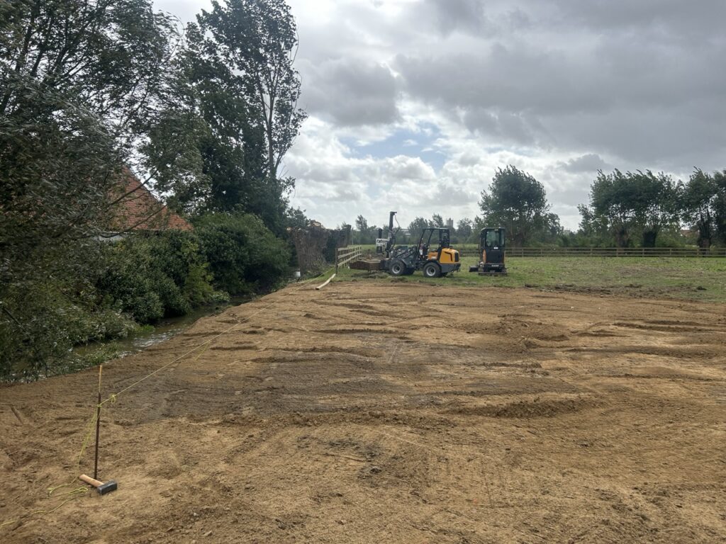 Nieuwe omheining wordt aangelegd met grondwerk en bouwmachines op open veld met bomen en waterloop
